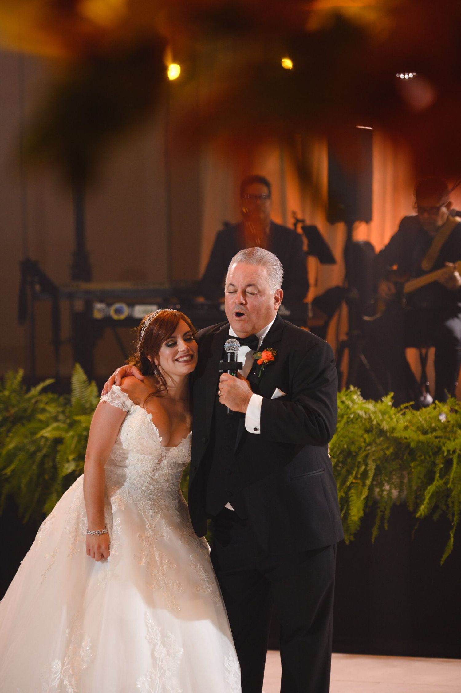 Father of the bride sings into microphone with daughter in white lace wedding gown at elegant reception.