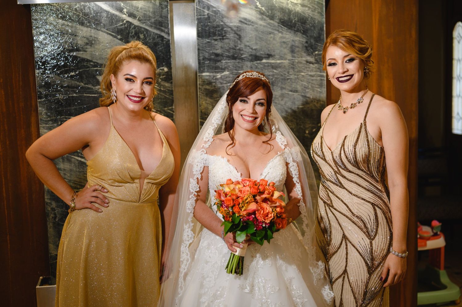 Bride in white lace gown with veil holds orange bouquet, flanked by two bridesmaids in gold dresses.