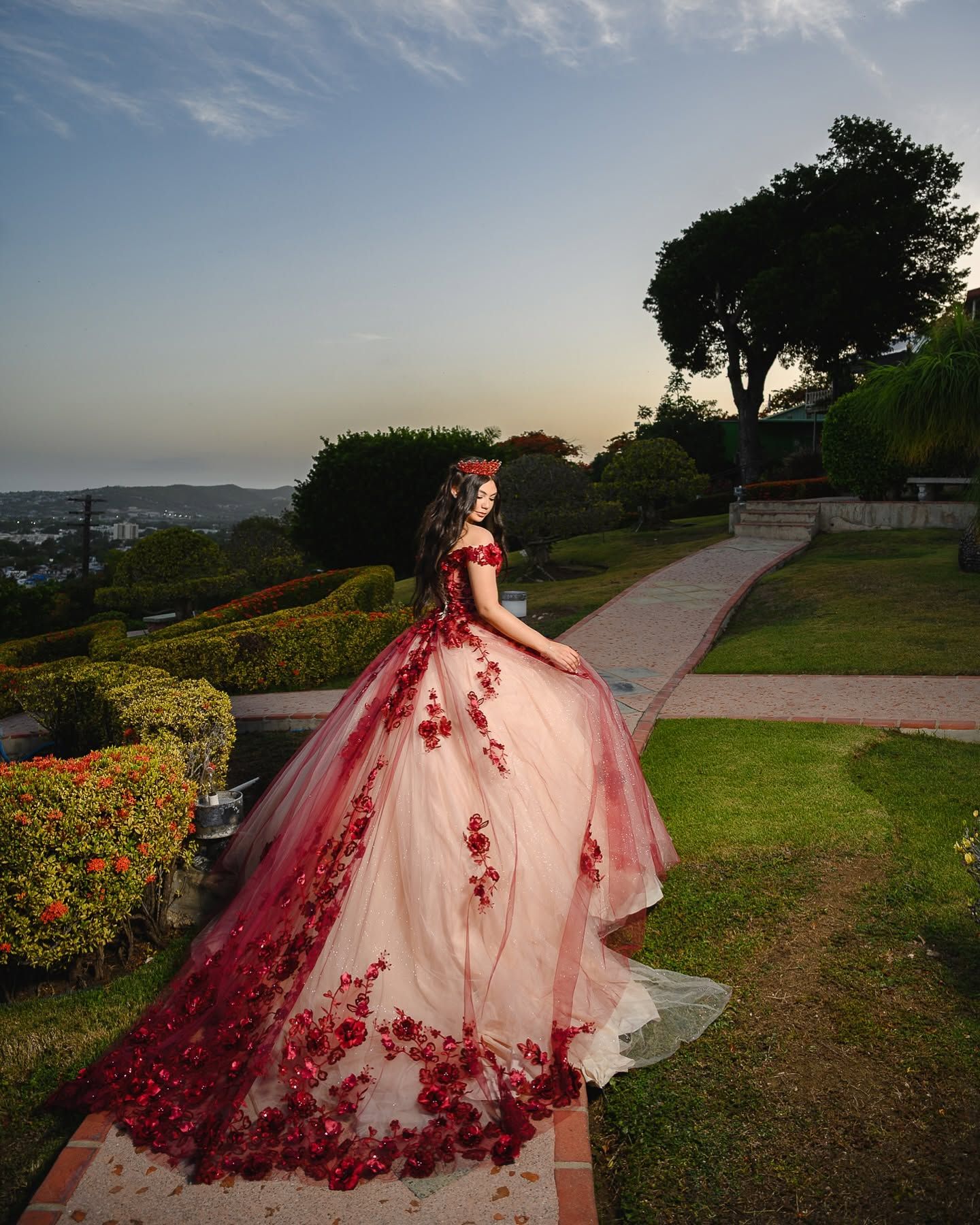 Woman in stunning red floral ball gown poses gracefully on garden path at sunset with panoramic city views.