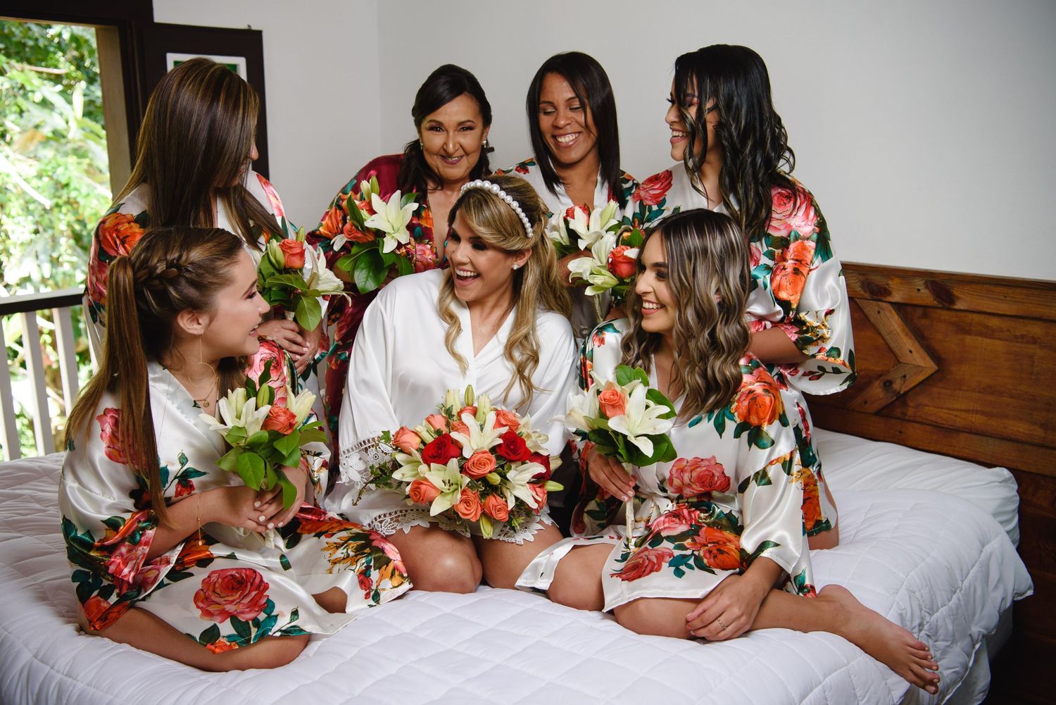 Bride in white robe with bridesmaids in floral robes holding colorful bouquets, laughing together on a bed.