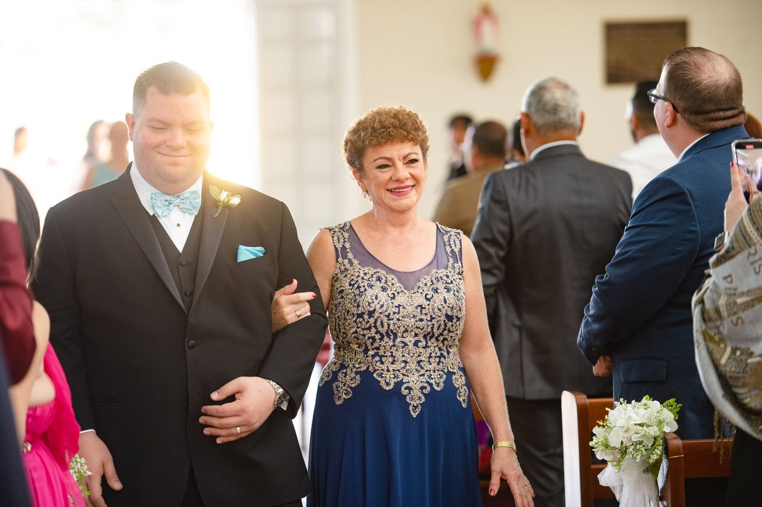 A groom in a black tuxedo walks down the church aisle with a woman in a blue embroidered gown during a wedding ceremony.