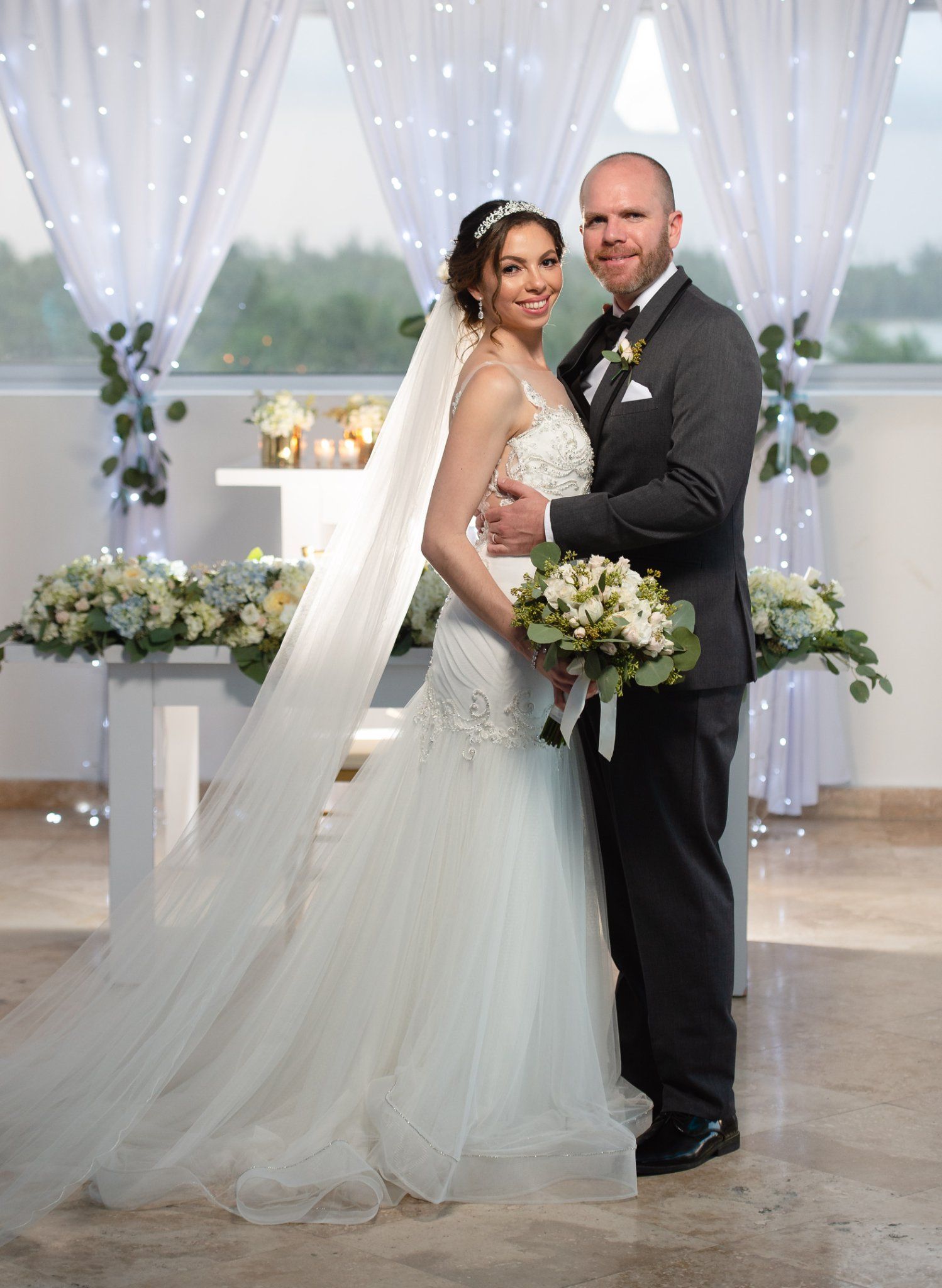 Bride in mermaid gown with veil and groom in dark suit pose together at elegant wedding ceremony venue.