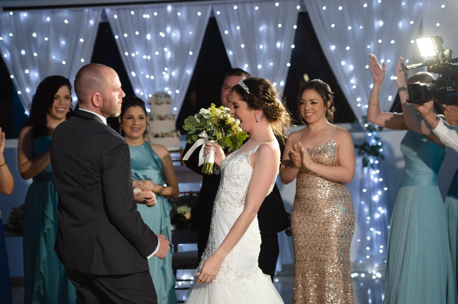 Bride and groom share a joyful wedding ceremony moment surrounded by bridesmaids in teal dresses against a sparkling backdrop.