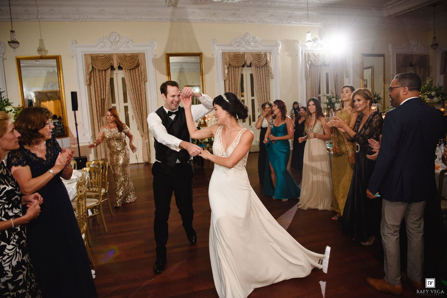 Bride and groom share first dance at elegant wedding reception with guests cheering in ornate ballroom.