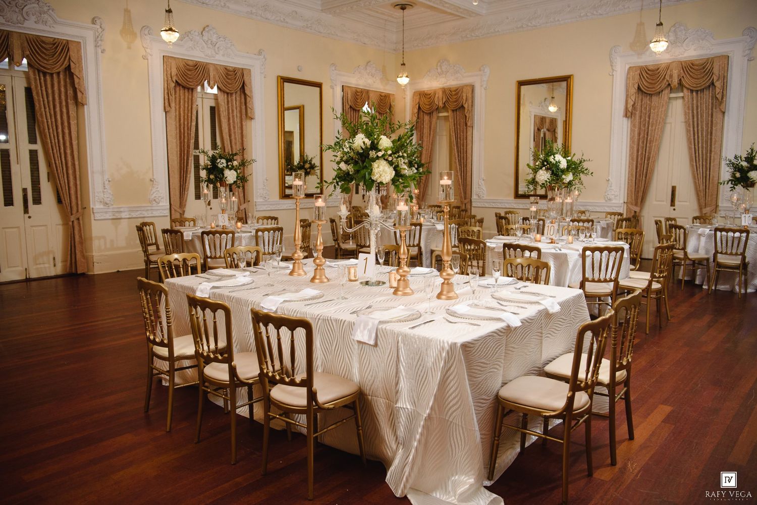 Elegant wedding reception ballroom with white tablecloths, gold chairs, tall floral centerpieces, and ornate decor.