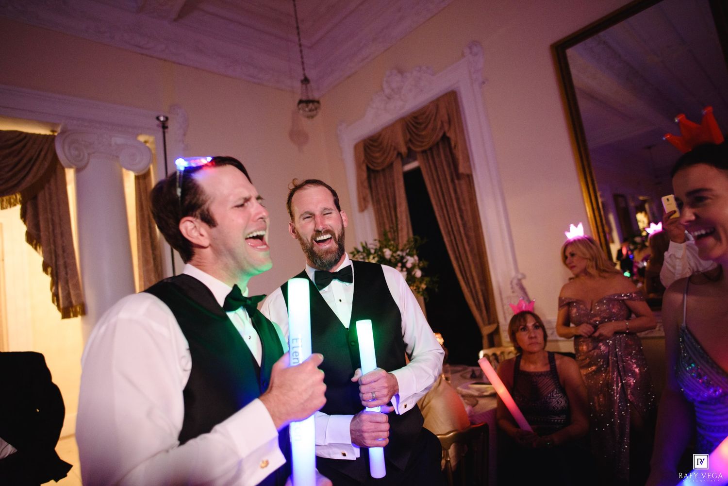 Two grooms laugh joyfully holding glow sticks at their wedding reception in an elegant venue.