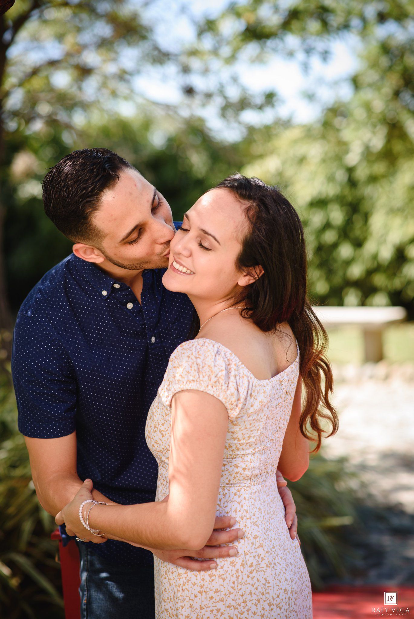A couple shares a romantic moment outdoors, man kissing womans cheek as she smiles, surrounded by lush greenery.
