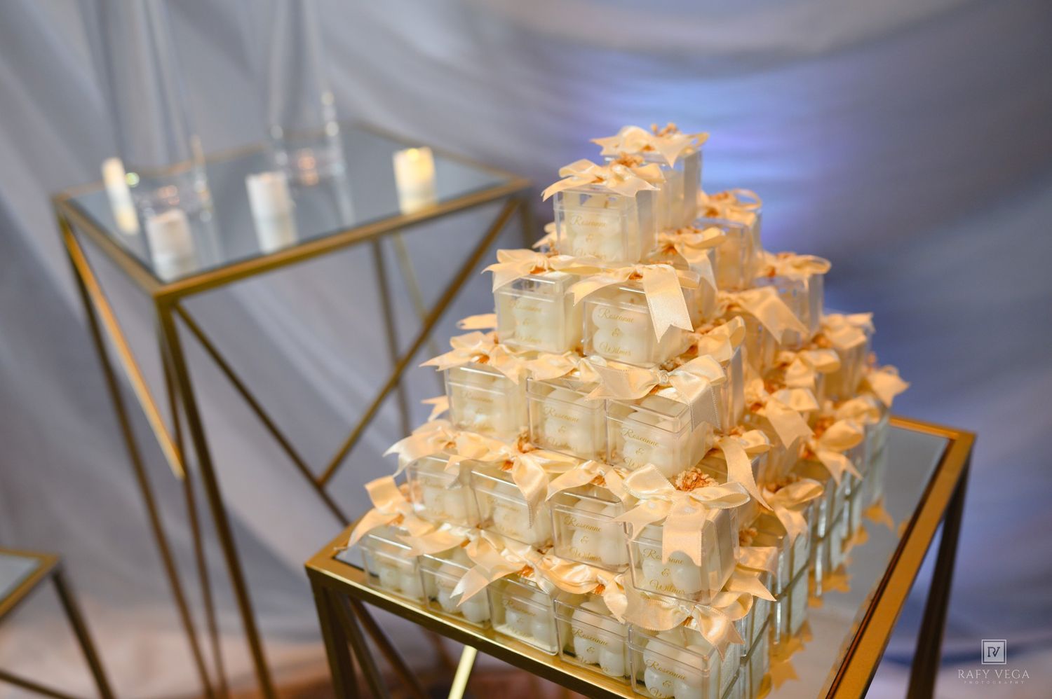 Elegant pyramid of white gift boxes with ribbons displayed on a gold geometric stand at a wedding reception.