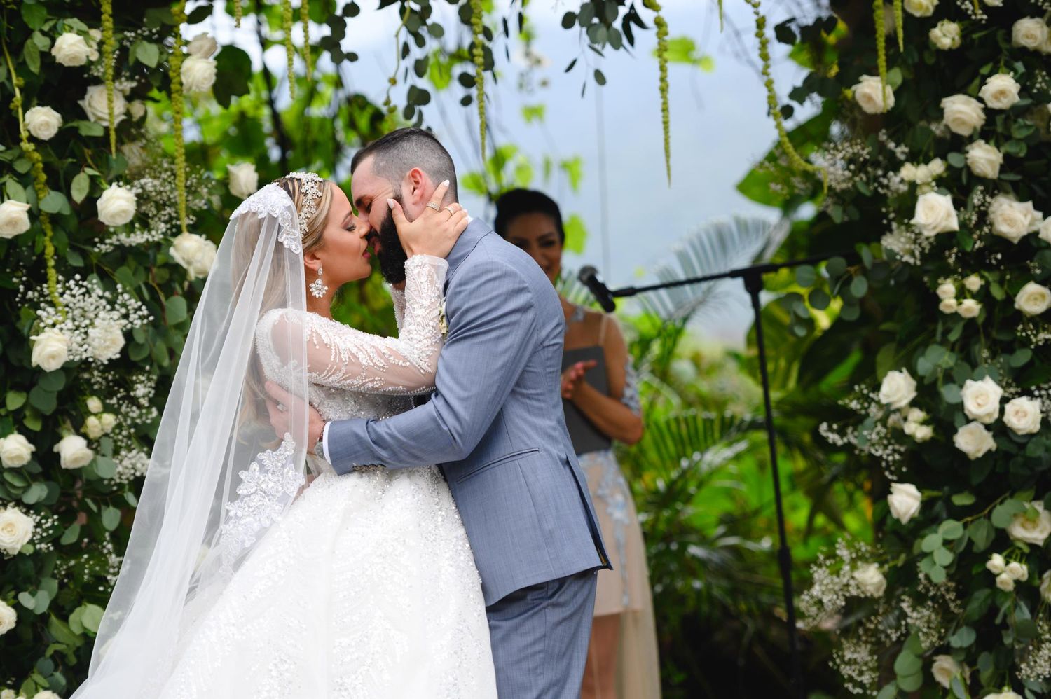 Bride and groom share first kiss at outdoor wedding ceremony beneath lush floral arch adorned with white roses and greenery.