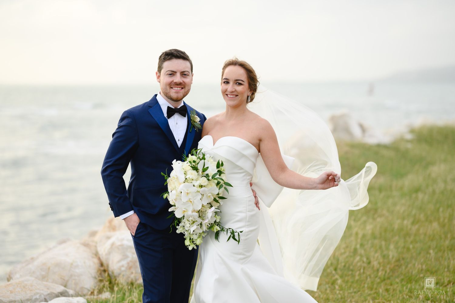Bride and groom pose by seaside, bride in strapless white gown with bouquet, groom in navy suit with bow tie.