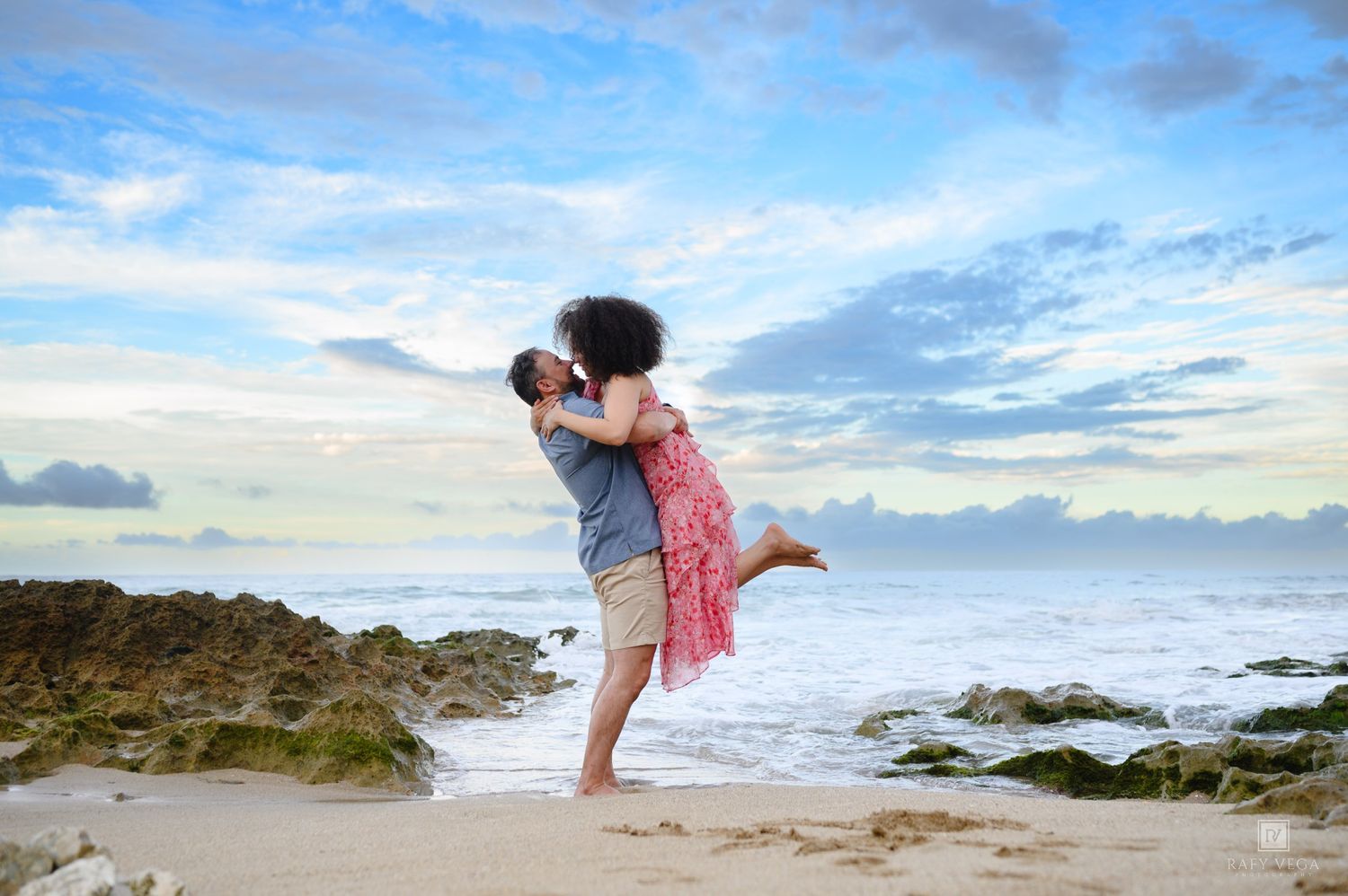 Couple embracing and kissing on a tropical beach with rocky shores and a blue cloudy sky at sunset.