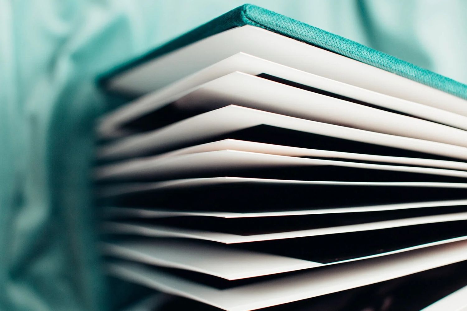 Close-up of a thick hardcover book with teal cover showing fanned white pages against a soft teal background.