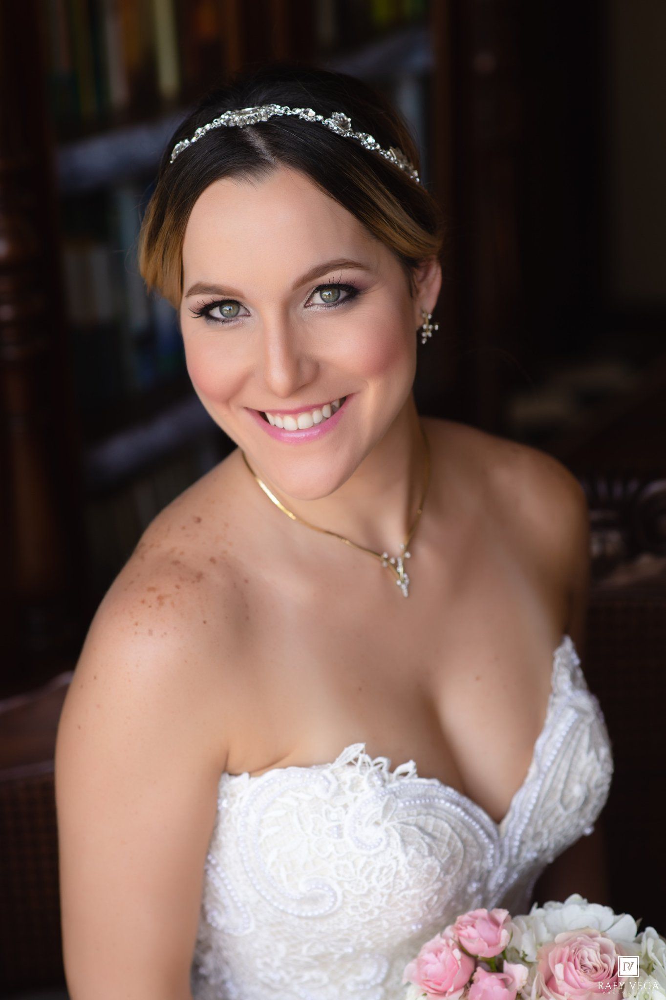 Smiling bride in strapless lace gown wearing crystal tiara and necklace, holding pink roses bouquet on her wedding day.