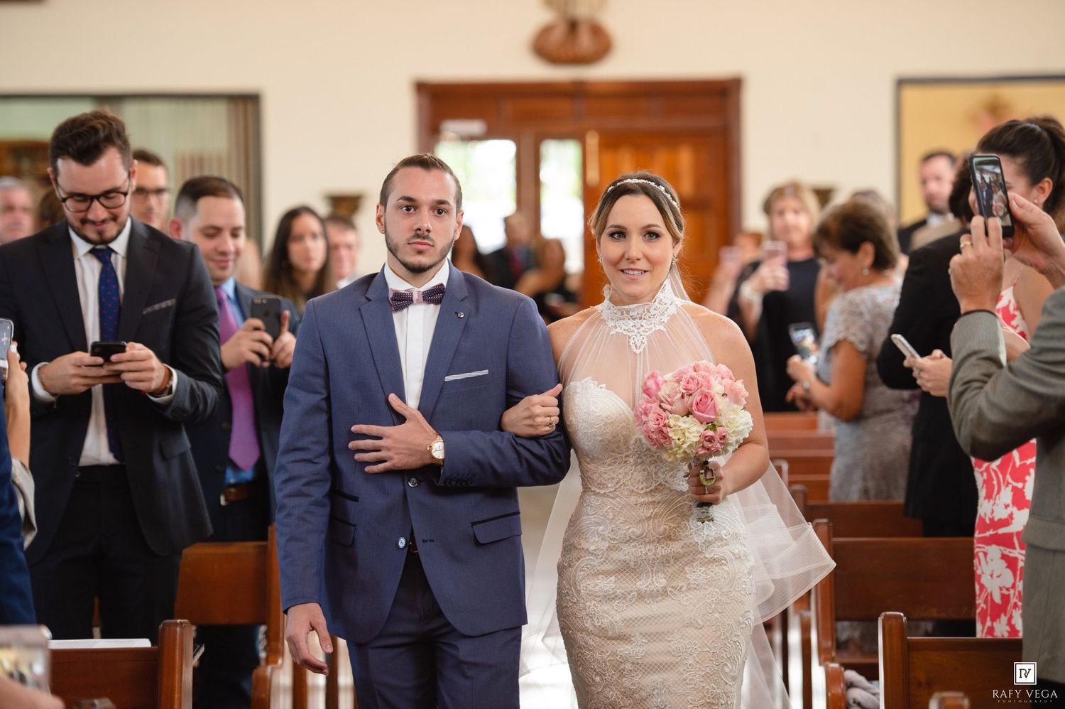 Bride and groom walking down church aisle together, surrounded by smiling wedding guests, bride holding white bouquet.