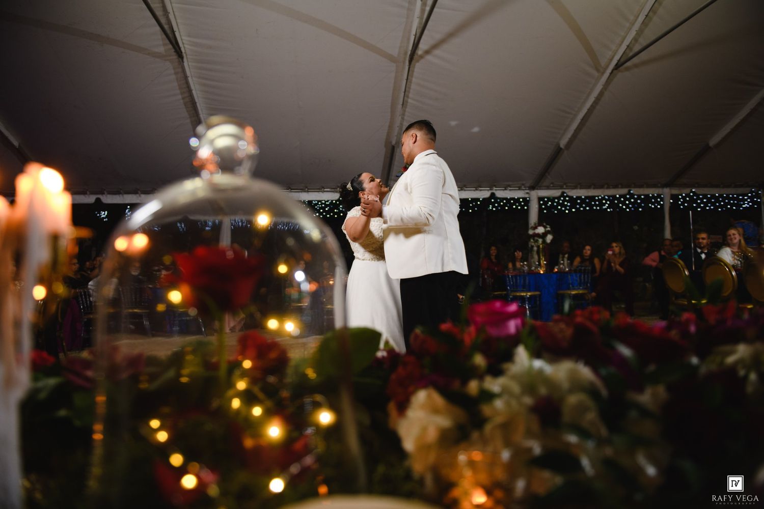 Bride and groom share first dance under white tent at elegant evening wedding reception with floral decorations.