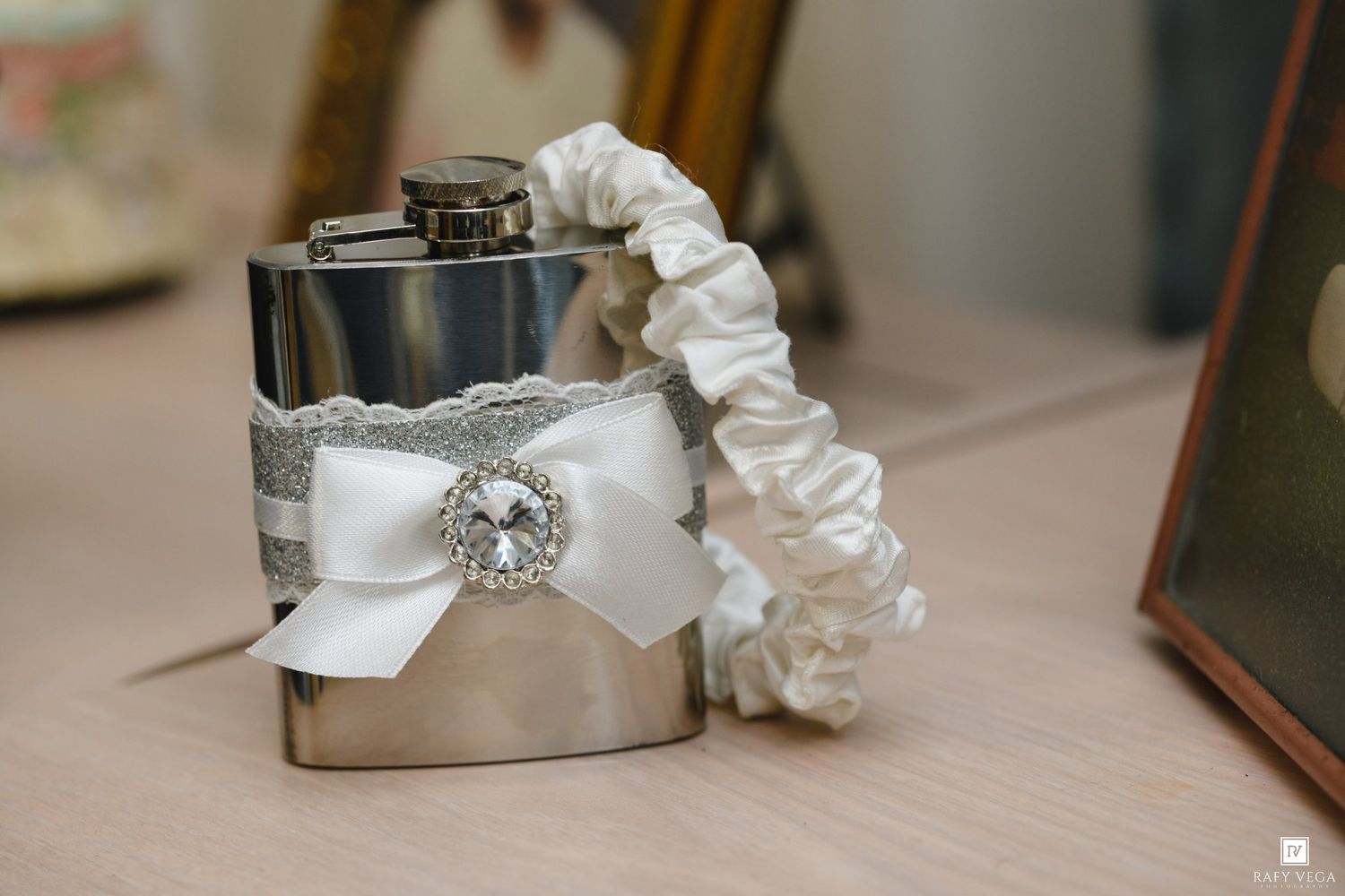 Silver hip flask decorated with lace, ribbon, and rhinestone brooch beside a white satin wedding garter on a wooden surface.