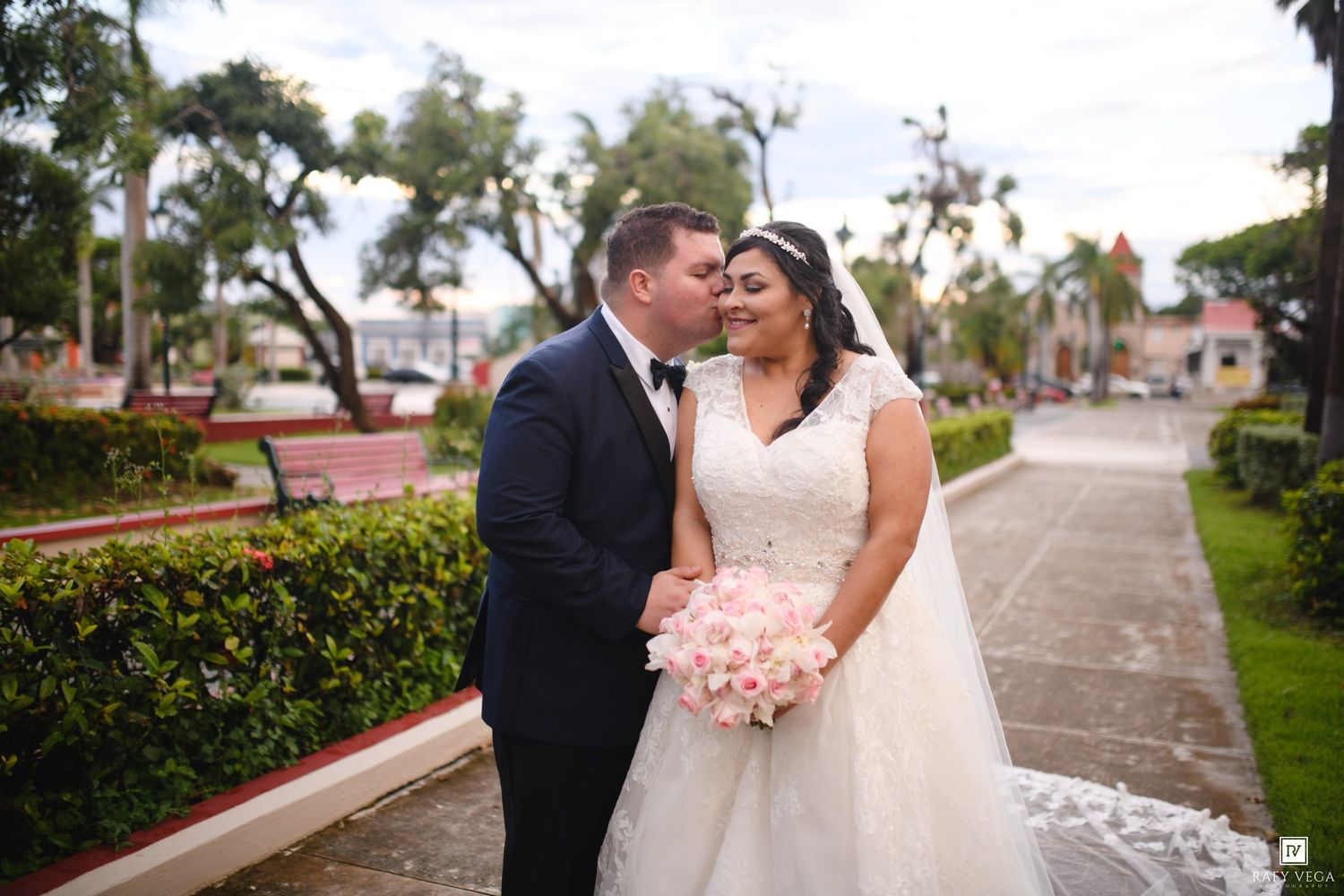 A groom in a navy suit kisses his bride in a white lace gown holding pink roses in a lush garden setting.