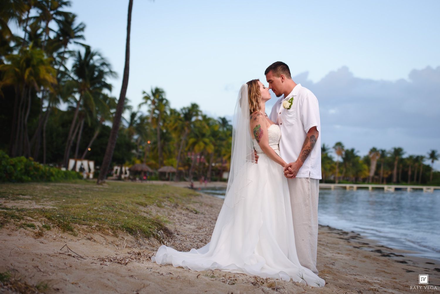 Bride and groom share romantic beach moment at sunset, surrounded by tropical palm trees near a serene ocean pier.