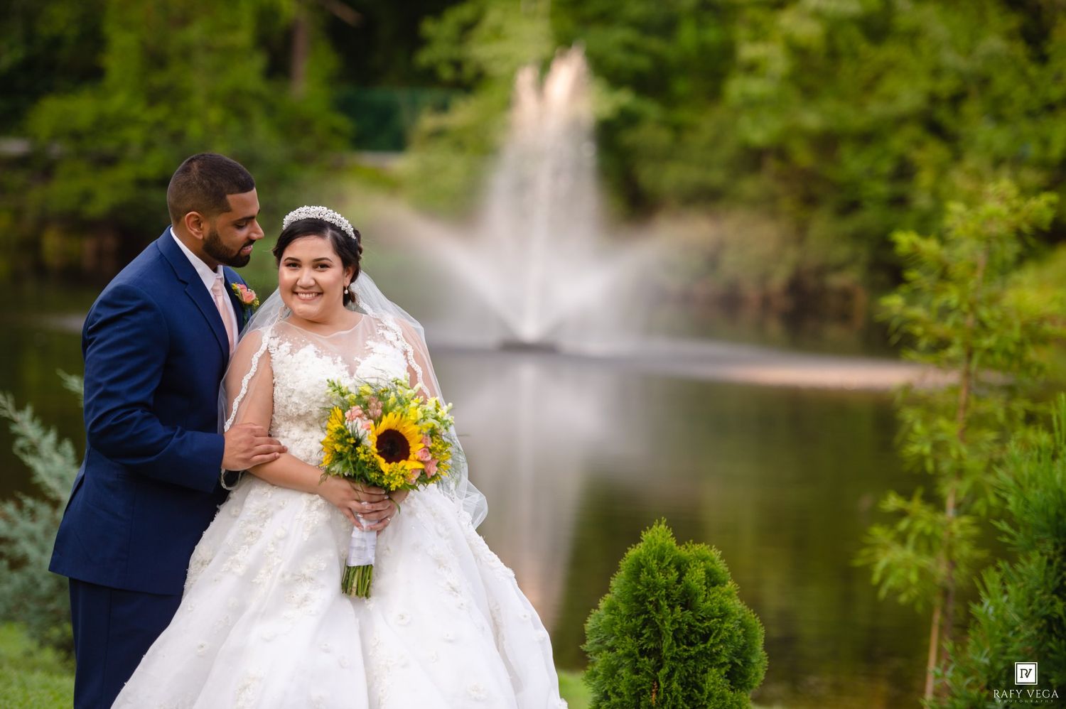 Bride in white ballgown holding sunflower bouquet poses with groom in navy suit near fountain in lush garden.