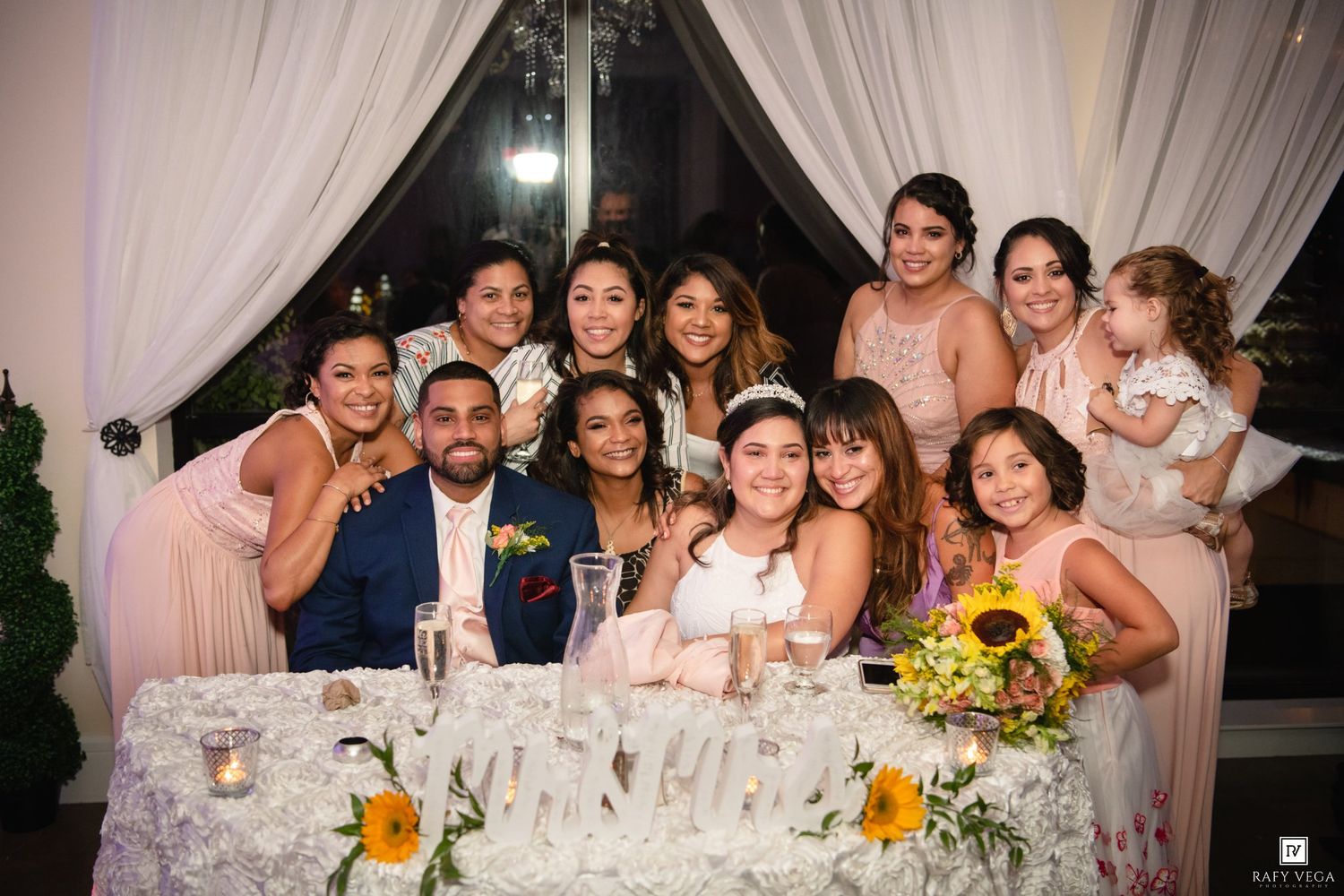 Wedding party poses at decorated sweetheart table with bridesmaids in blush gowns, flower girls, and sunflower centerpieces.
