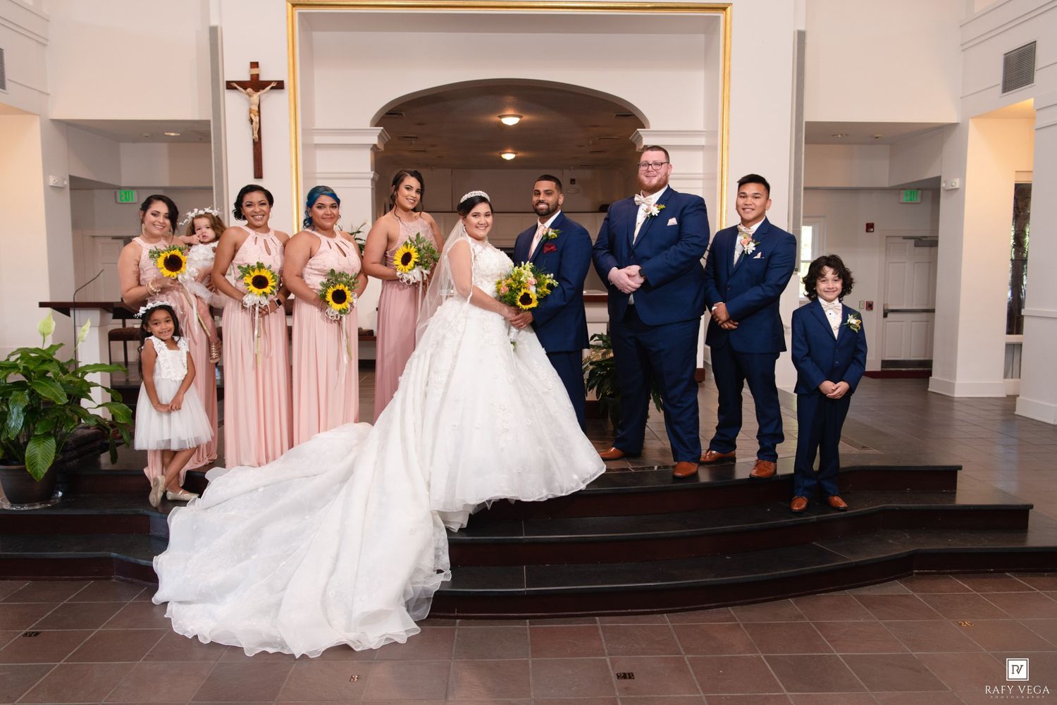 Wedding party poses in a church, bride in white gown with bridesmaids in blush pink holding sunflowers and groomsmen in navy suits.