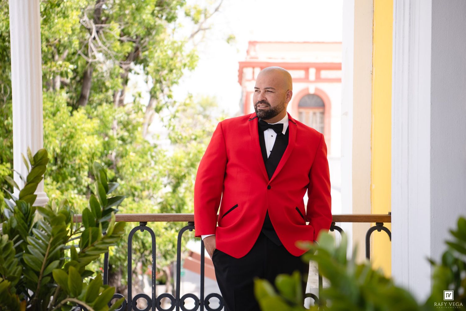 Man in red tuxedo jacket and black bow tie standing on a balcony surrounded by lush greenery.