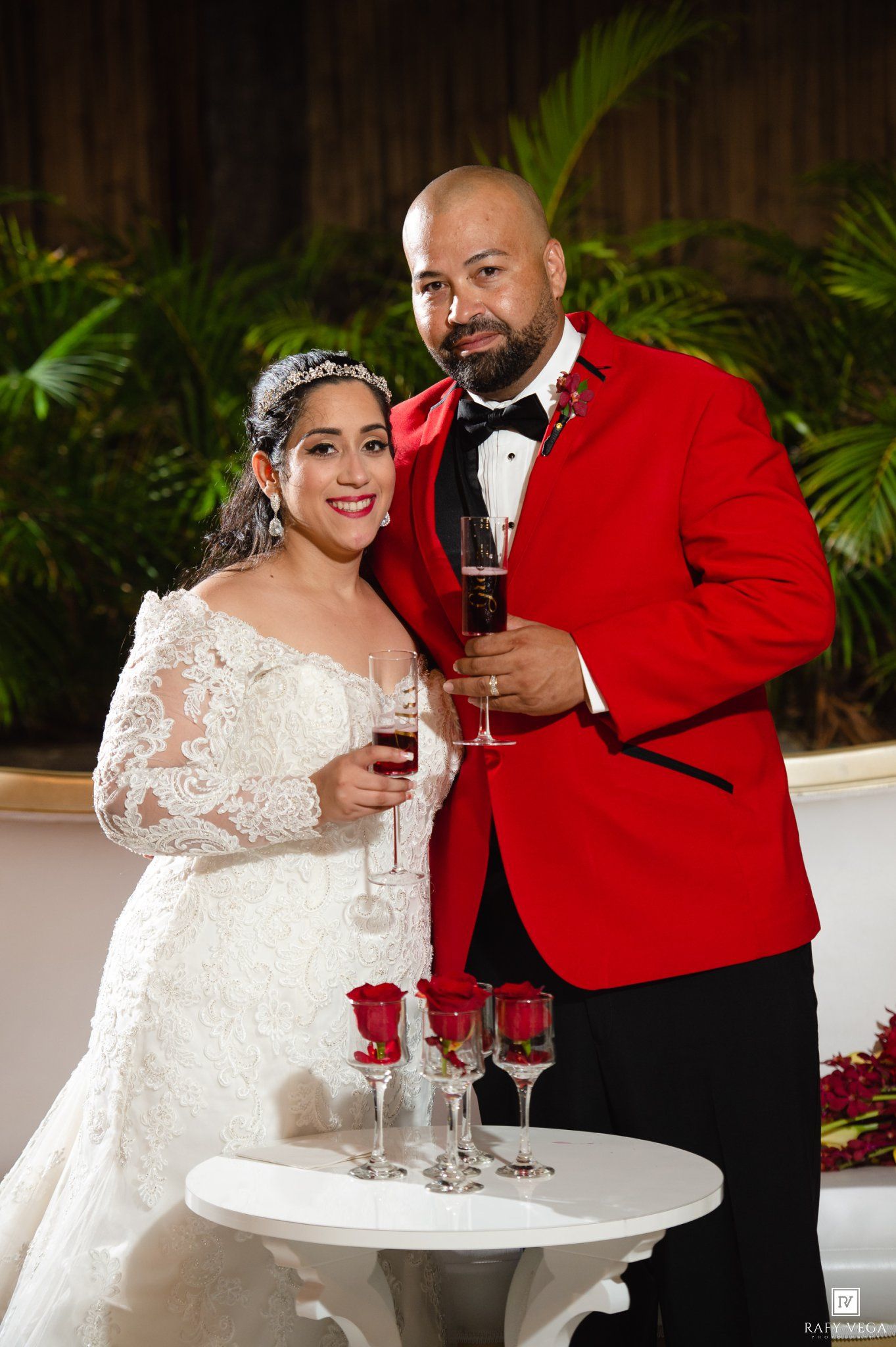 Bride in white lace gown and groom in red tuxedo jacket toasting with wine glasses at elegant wedding reception.