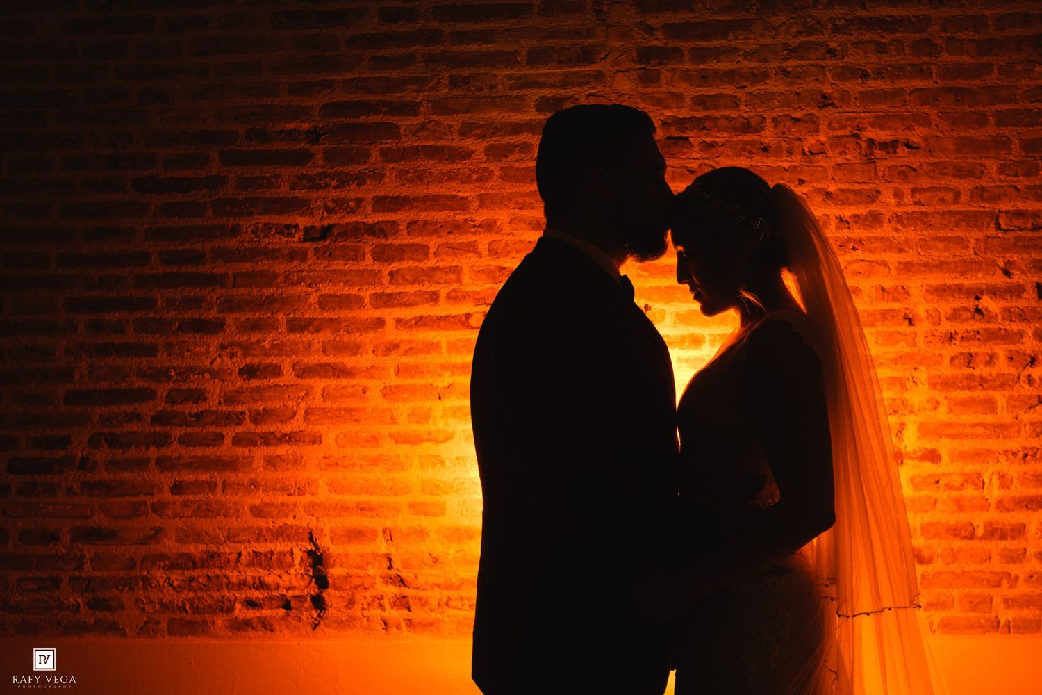 Silhouette of bride and groom sharing romantic moment against glowing orange brick wall.