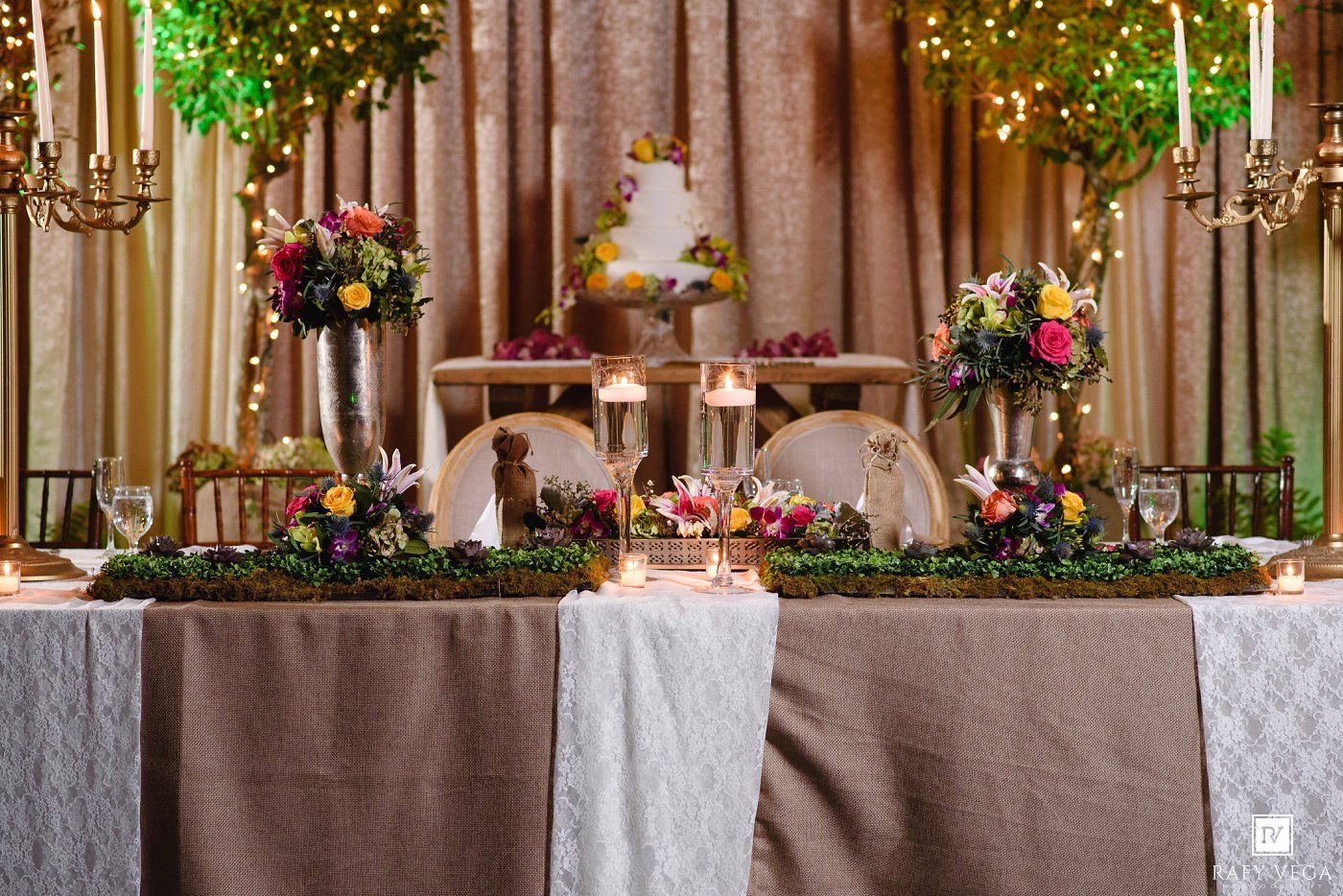 Elegant wedding head table with colorful floral arrangements, moss garland, candelabras, and a tiered cake on a draped table.