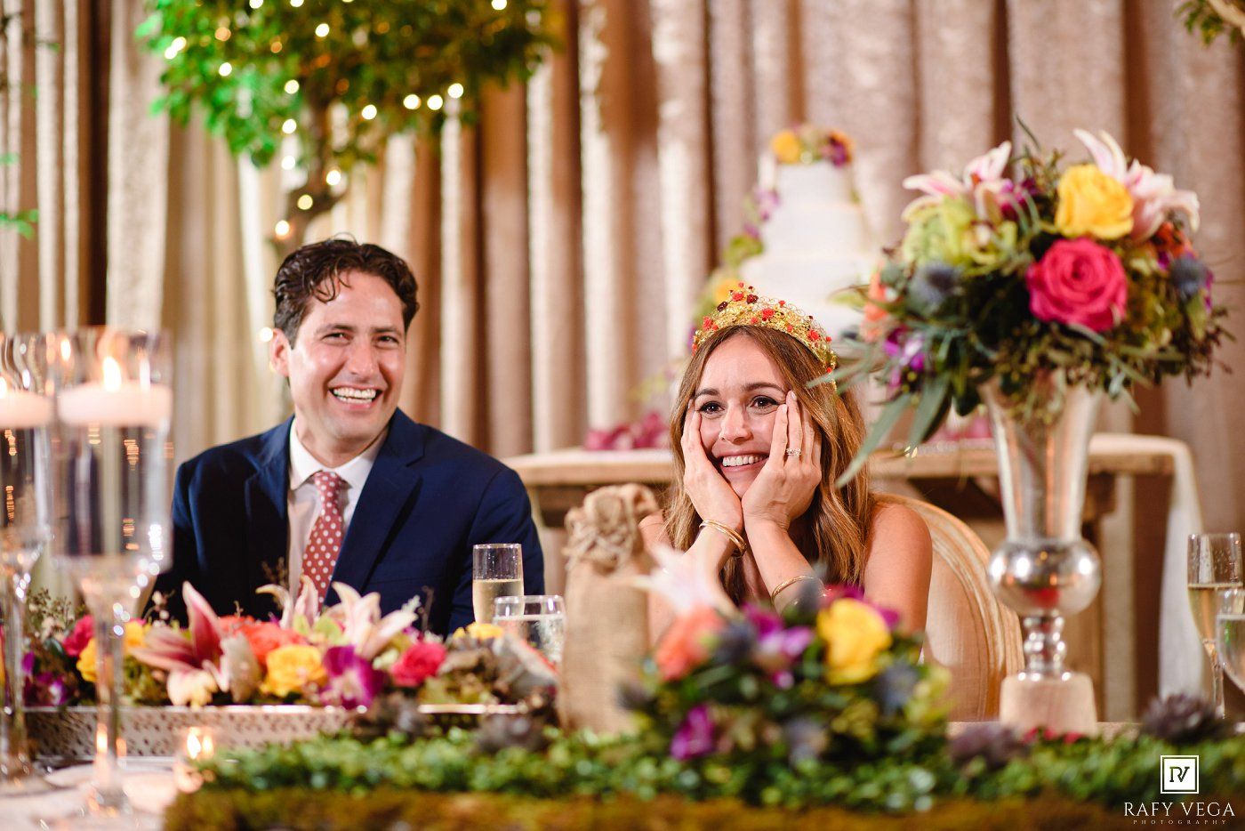 Smiling bride and groom seated at sweetheart table with colorful floral arrangements and wedding cake behind them.