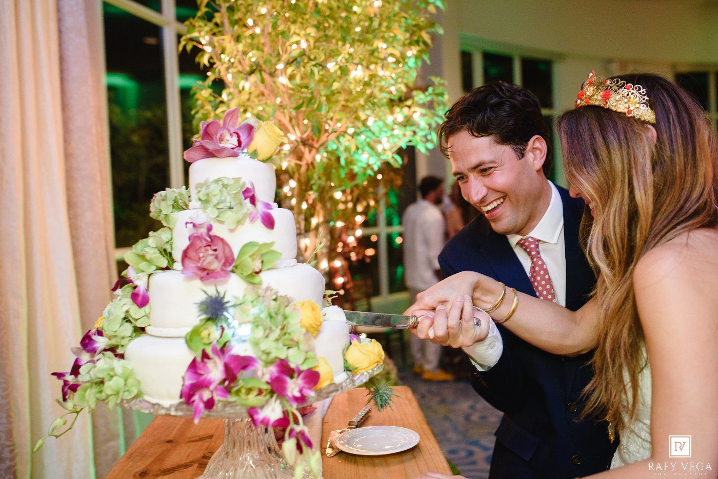 Couple cutting a floral wedding cake decorated with orchids and greenery at an elegant outdoor reception.