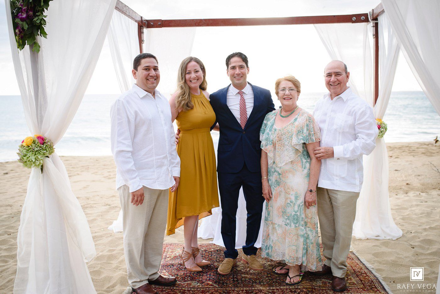 Five people pose together at a beach wedding ceremony under a decorated arch with white draping.