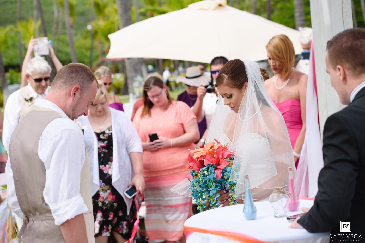 Bride in white gown and veil holds colorful bouquet during outdoor wedding ceremony with guests gathered around.