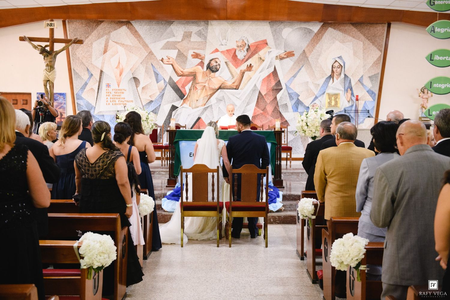 Couple kneeling at church altar during wedding ceremony with colorful religious mural backdrop and guests seated in pews.