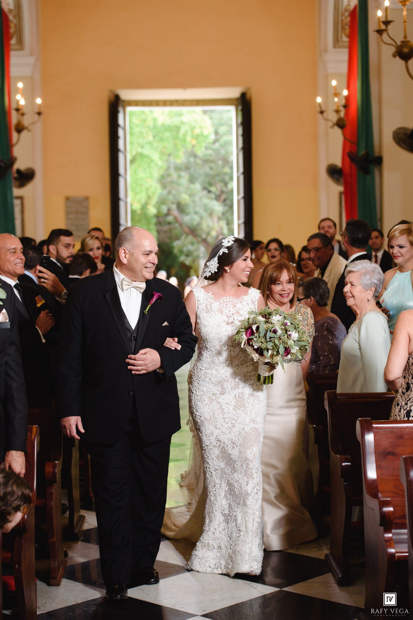 Bride in lace gown walks down church aisle with parents during elegant wedding ceremony.