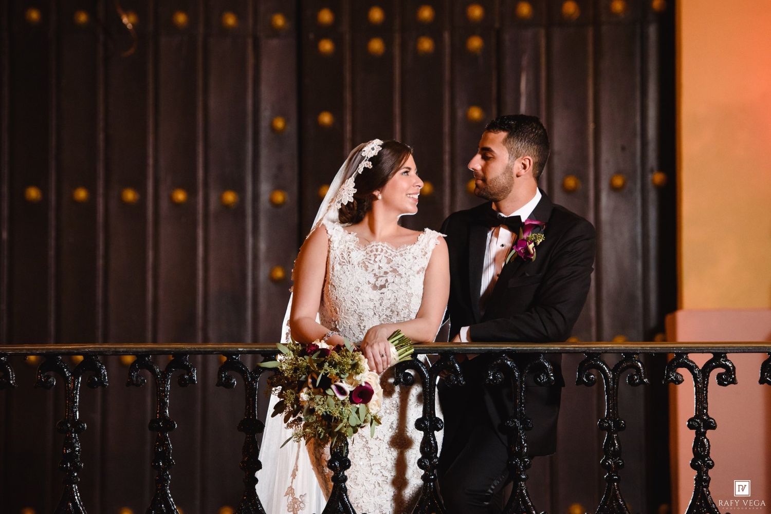 Bride and groom share romantic moment on ornate balcony, bride holding lush bouquet in elegant lace gown.