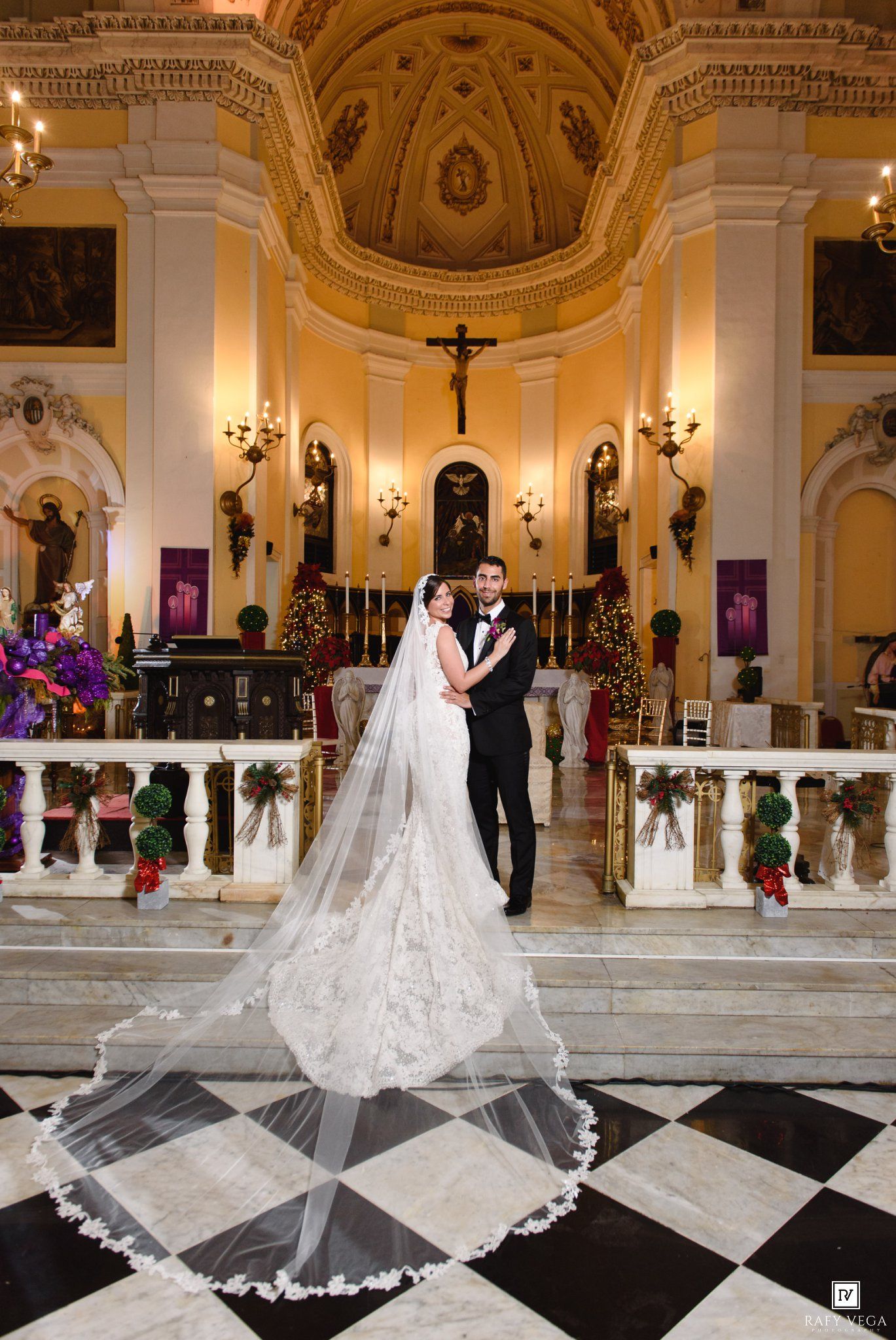 Bride and groom pose in elegant church with marble floors, ornate gold decor, and a long lace cathedral veil trailing behind.