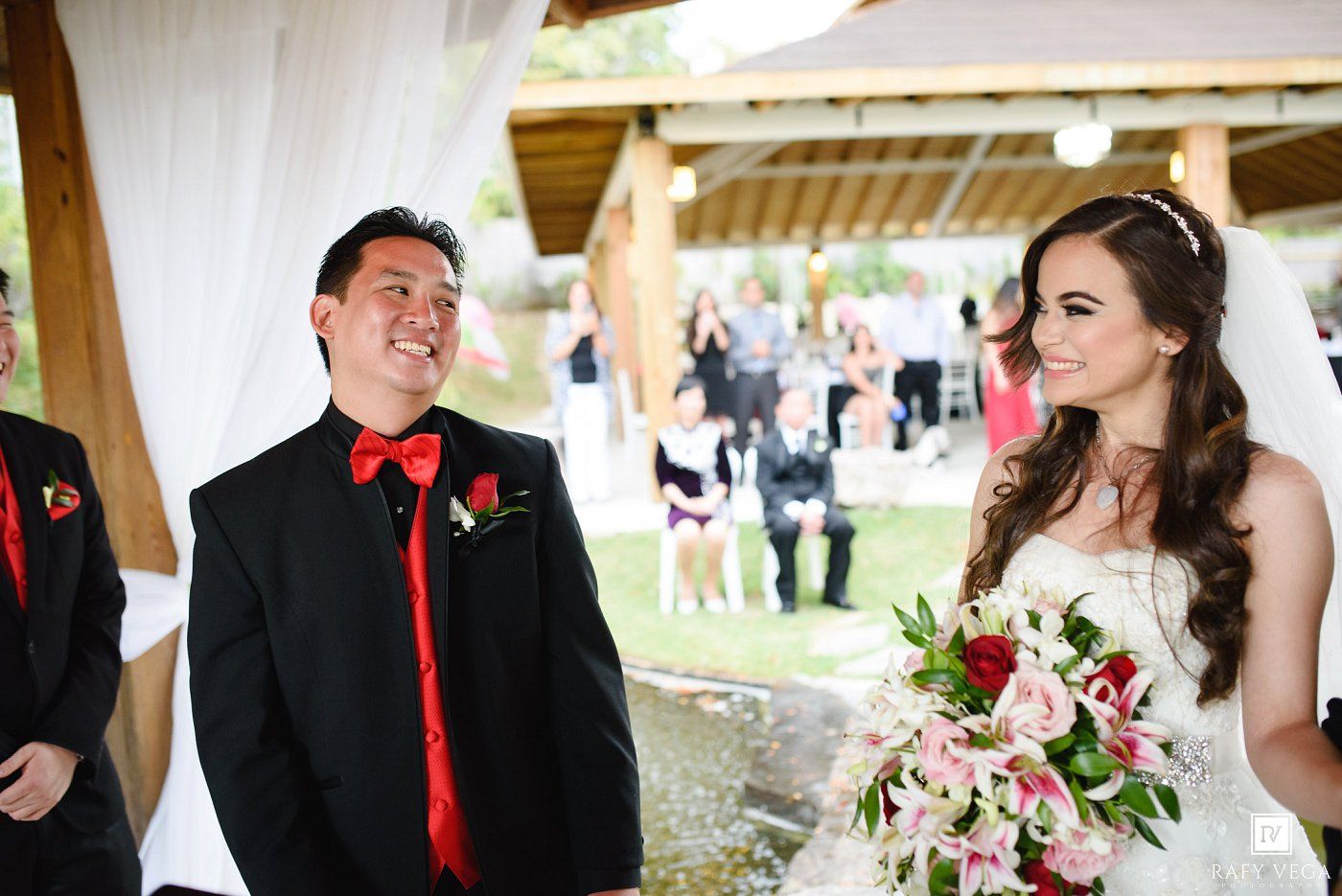 Groom in black tuxedo with red tie smiles at bride holding pink bouquet during outdoor wedding ceremony.
