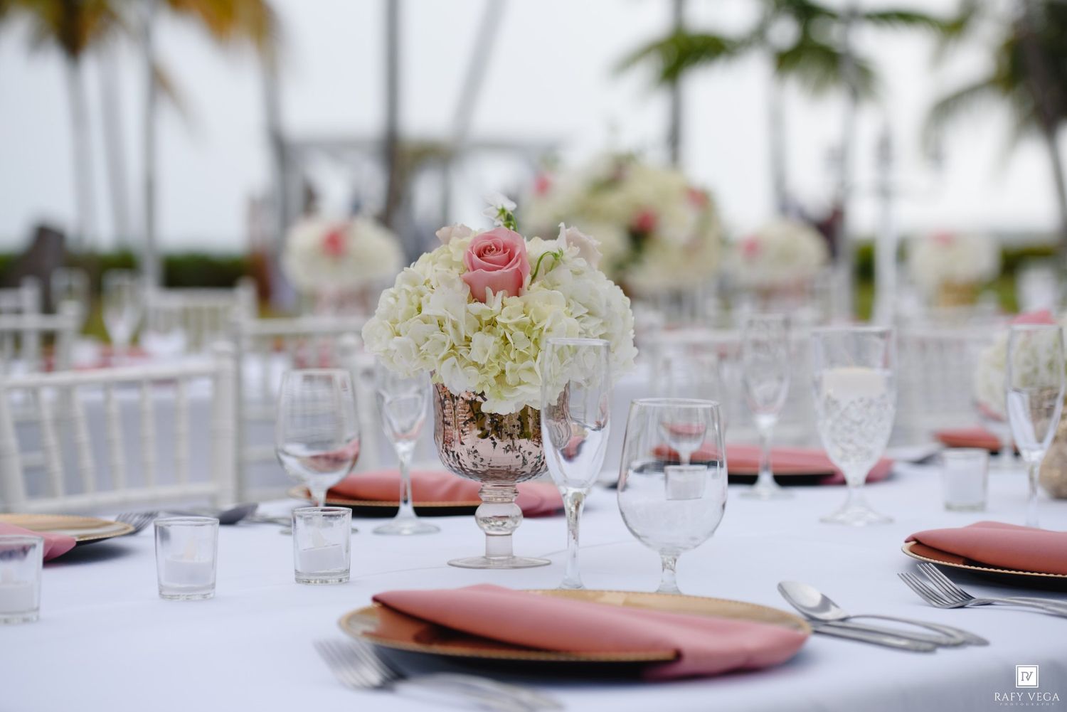 Elegant outdoor wedding reception table with white hydrangea and pink rose centerpiece, coral napkins, and crystal glassware.