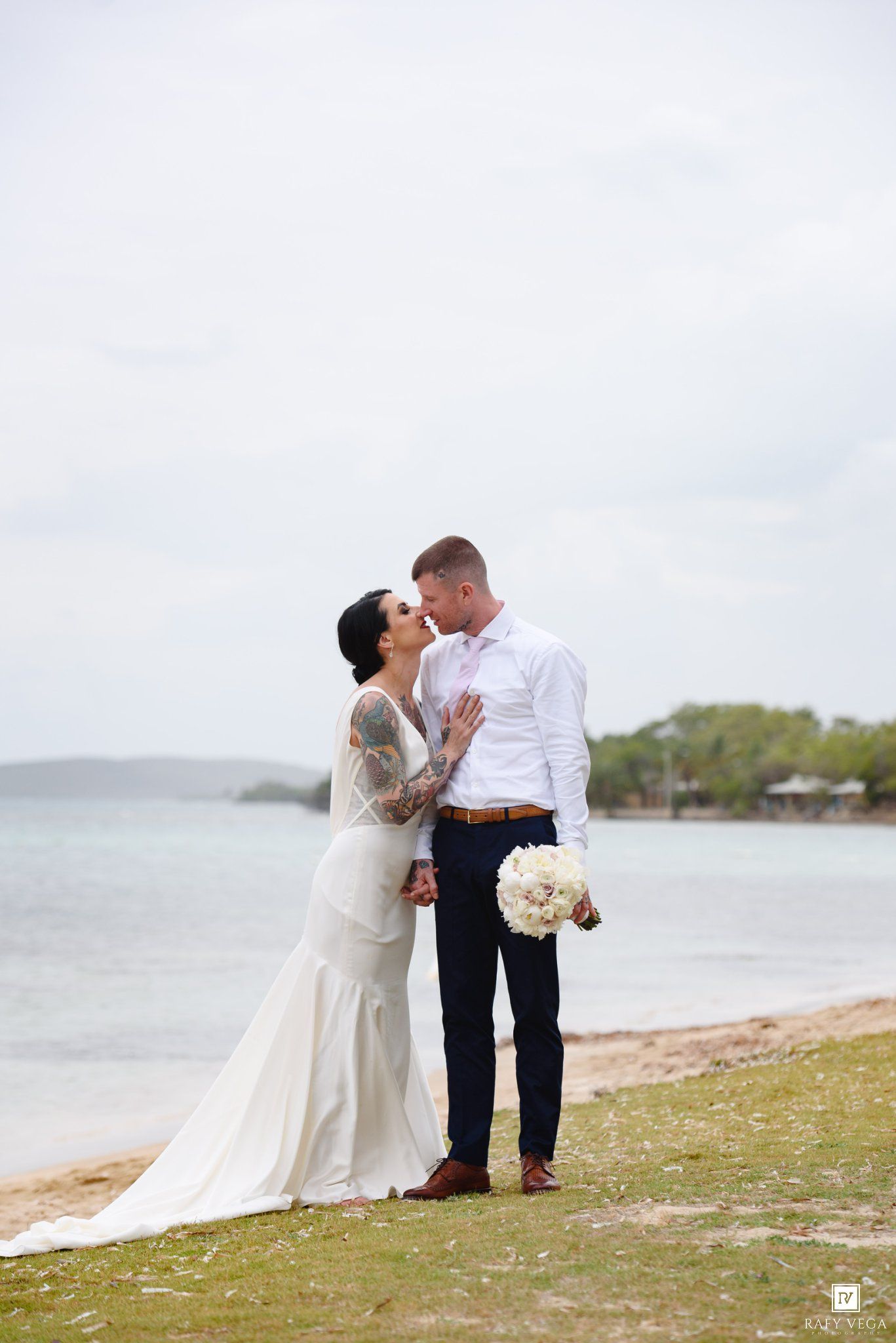 Bride and groom share a romantic moment on a beach, she in a white gown with tattoos, he in navy suit holding bouquet.