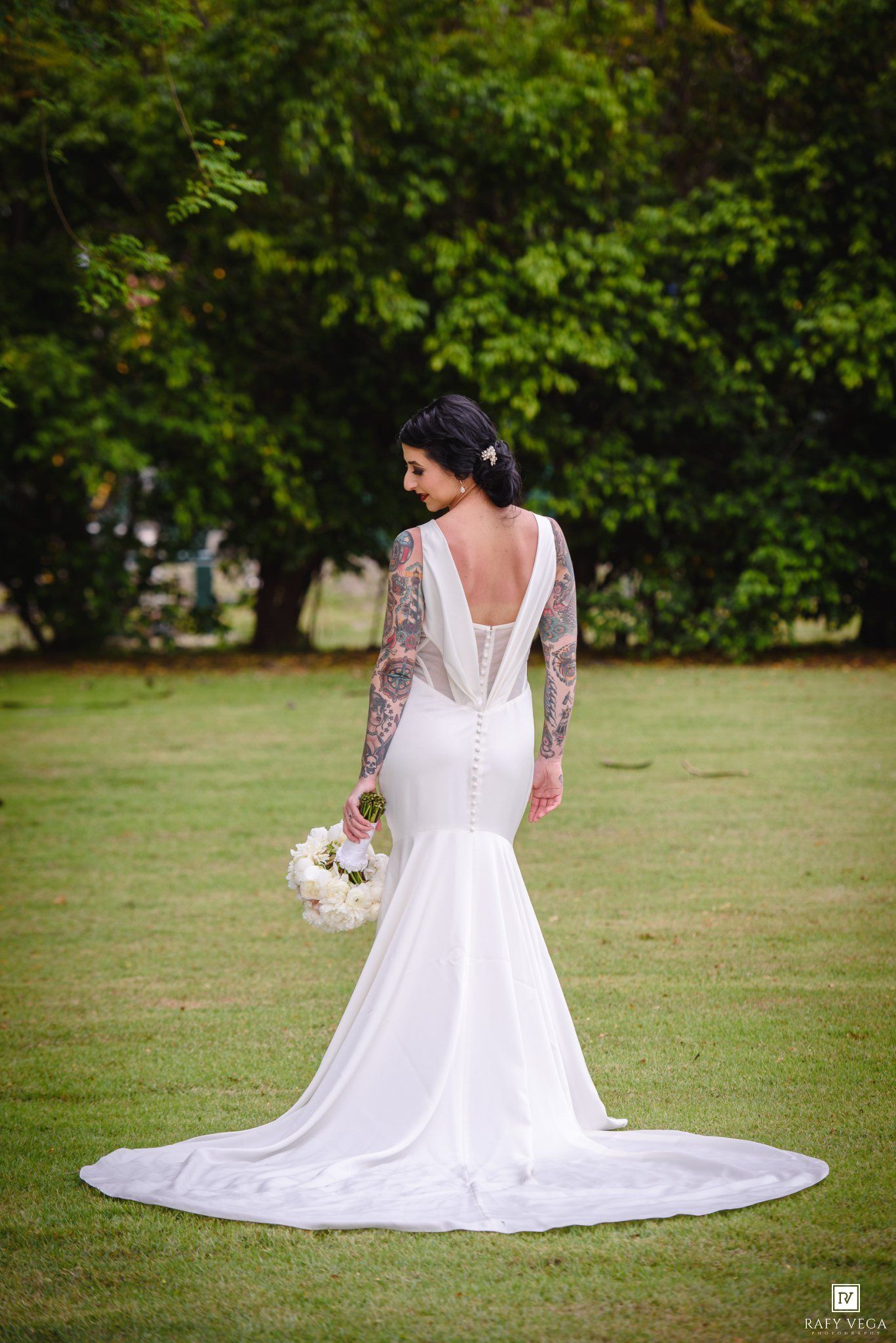 Bride in white mermaid wedding gown with lace sleeves and tattoos, holding bouquet, standing on green lawn.