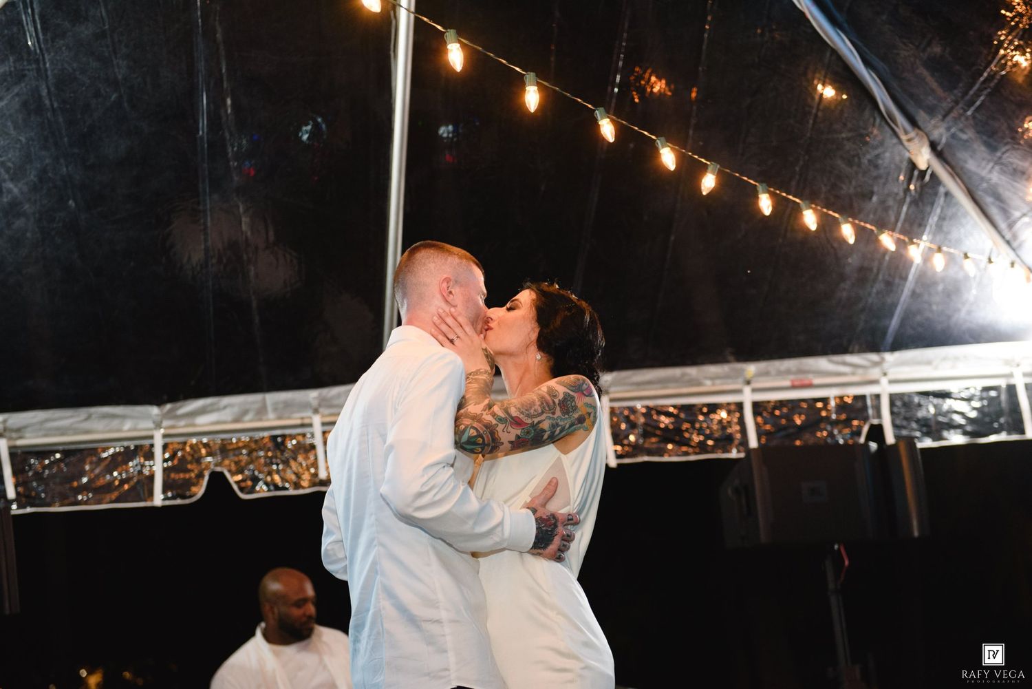 Tattooed bride and groom share a kiss under string lights in a wedding tent.