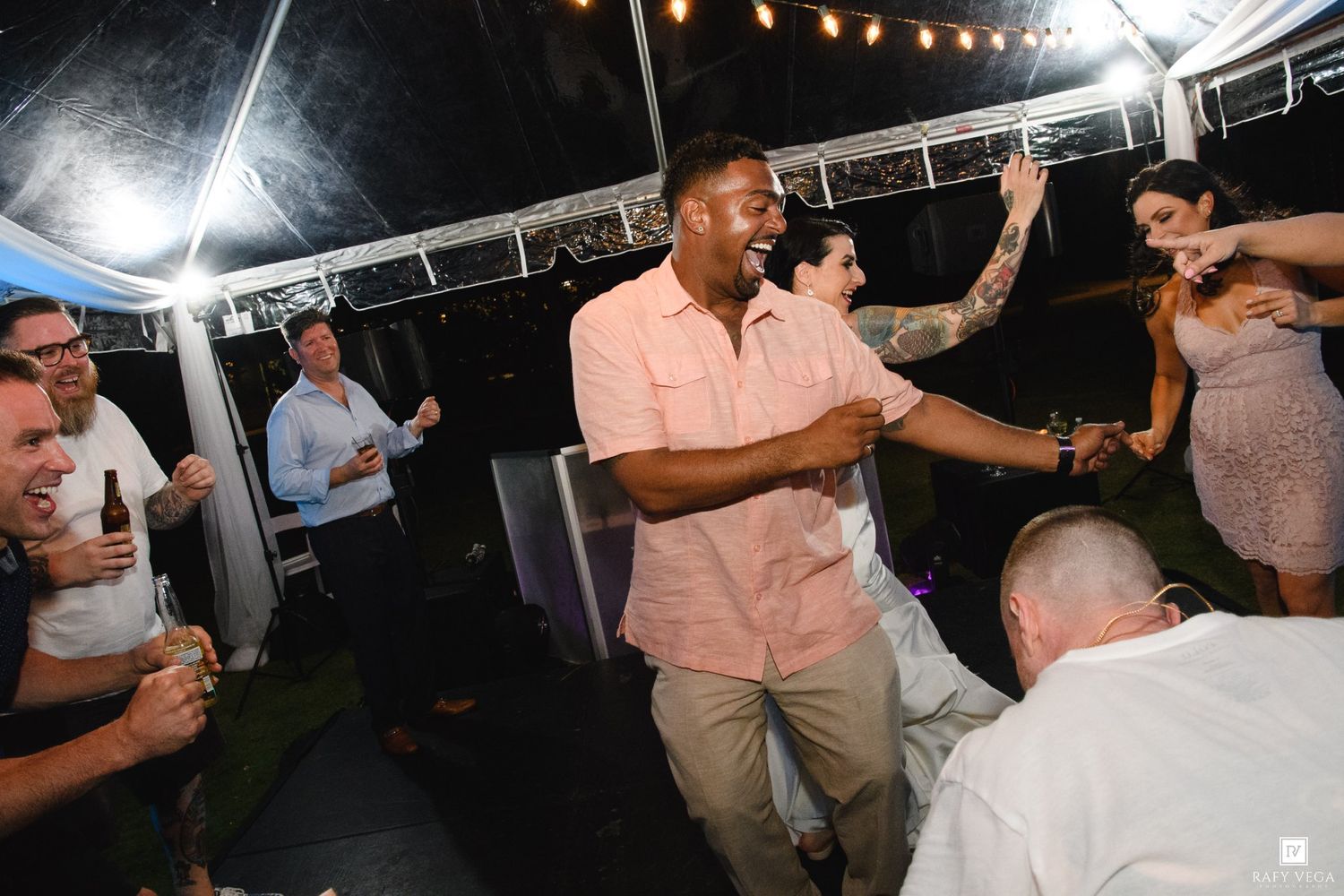 Guests dancing and celebrating at an outdoor evening wedding reception under a white tent with string lights.