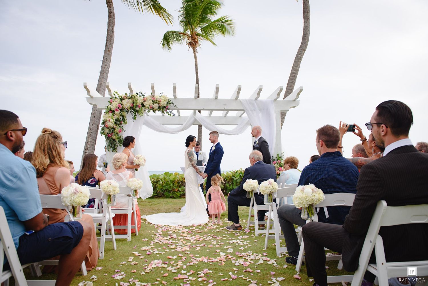 Outdoor beach wedding ceremony under floral arch with couple exchanging vows, guests seated on white chairs near palm trees.
