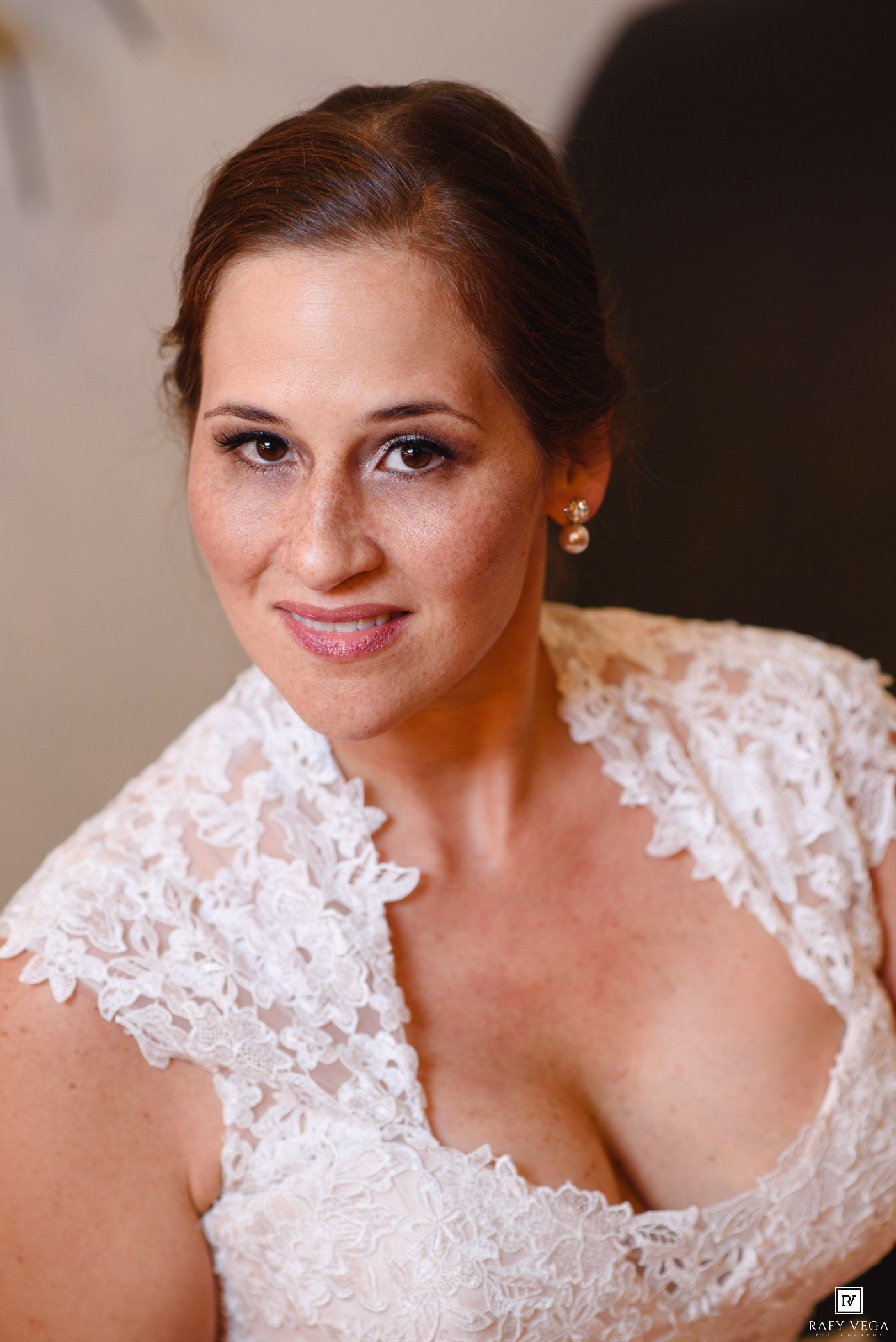 Close-up portrait of a bride in a white lace wedding dress, wearing pearl earrings, smiling softly.