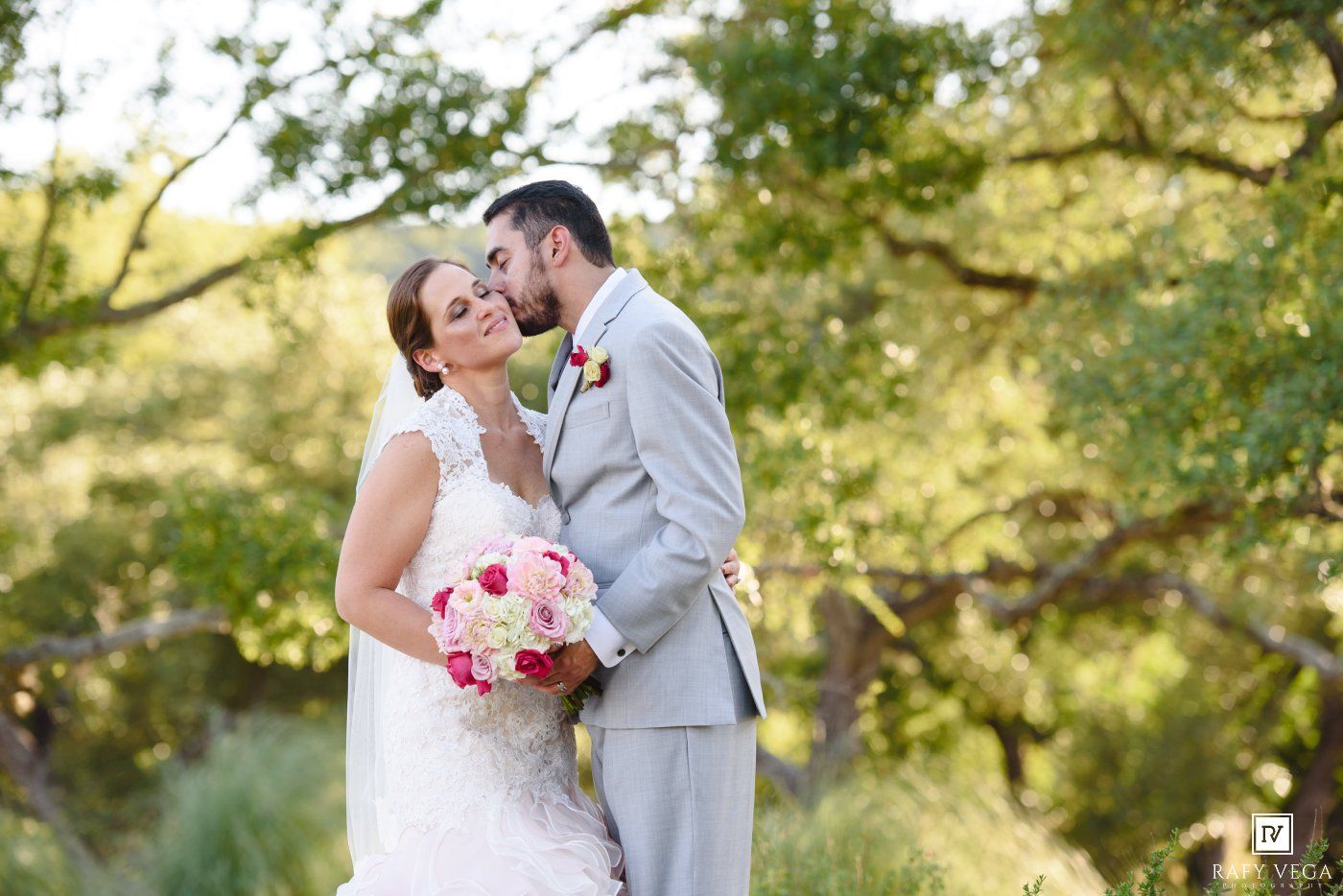 Bride and groom share a romantic kiss outdoors, surrounded by lush green trees, bride holding a pink and red bouquet.