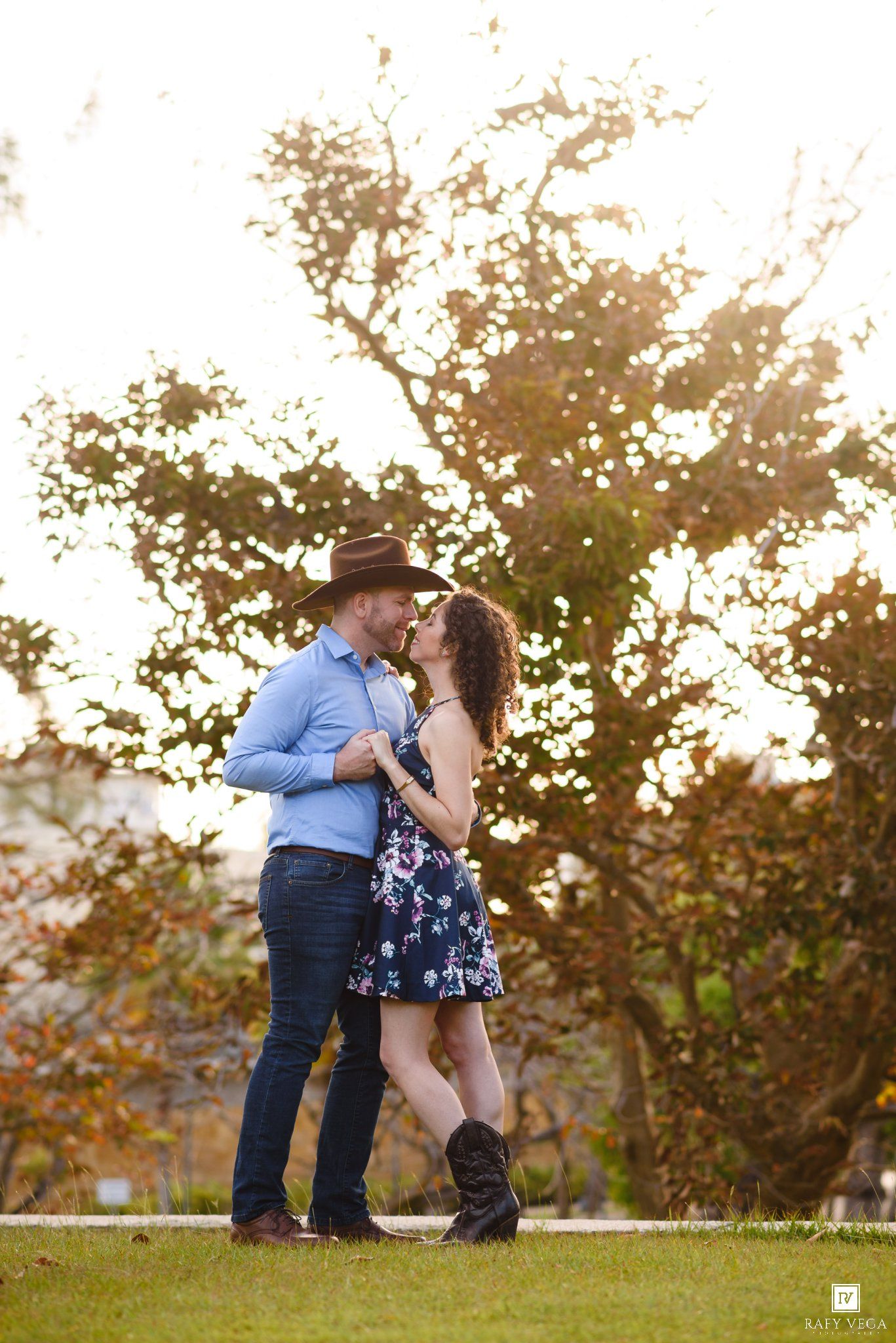 Couple sharing a romantic kiss outdoors, man in cowboy hat and blue shirt, woman in floral dress and boots, golden sunlight.