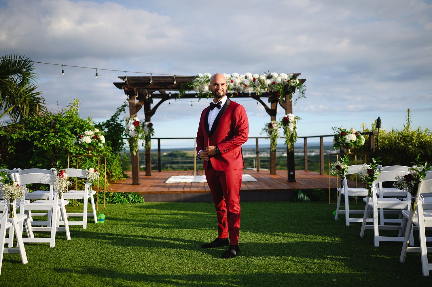 Groom in red tuxedo stands at outdoor wedding ceremony with floral arch and white chairs on green lawn.