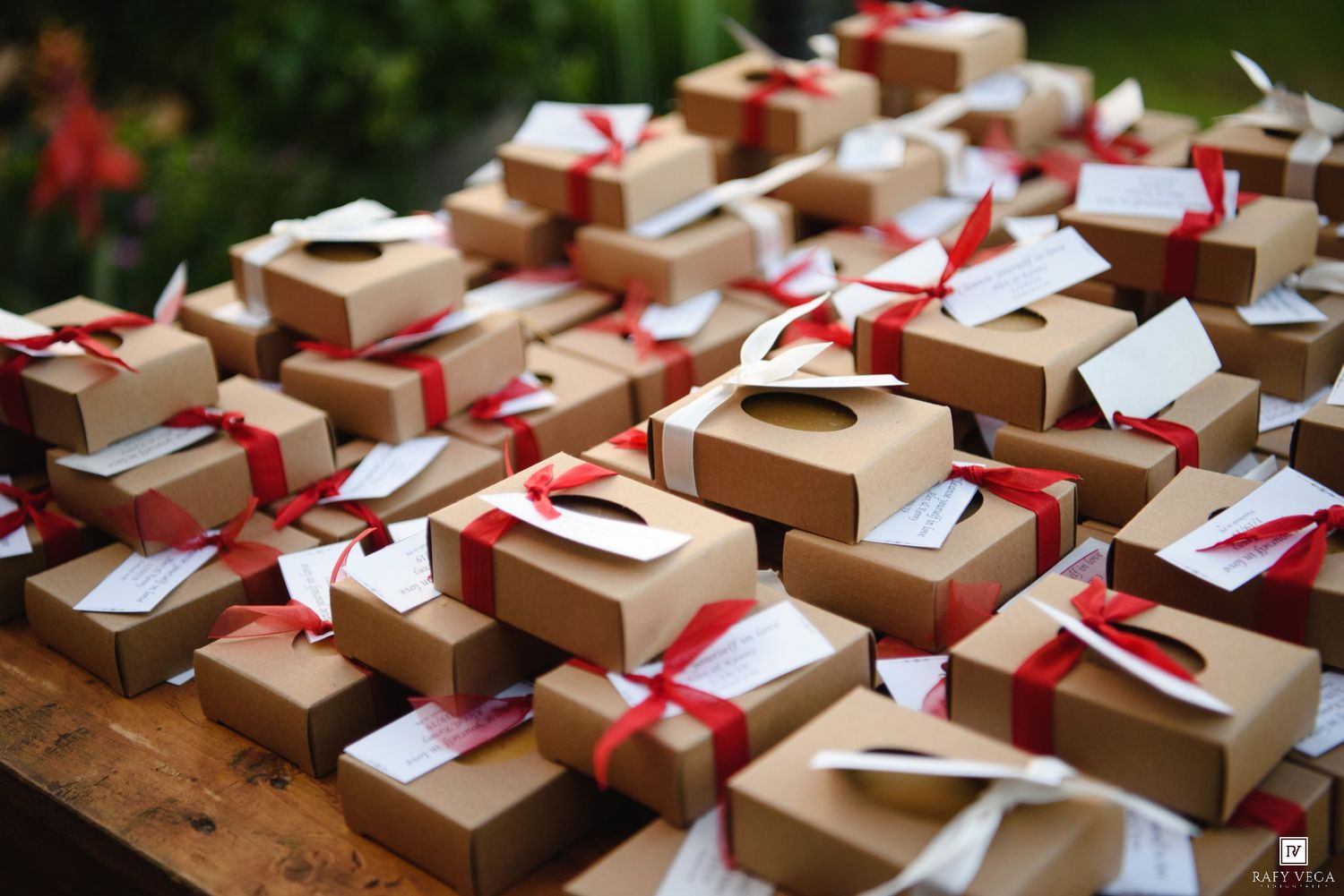 Brown kraft gift boxes tied with red ribbons and white tags displayed on a wooden table at an outdoor event.
