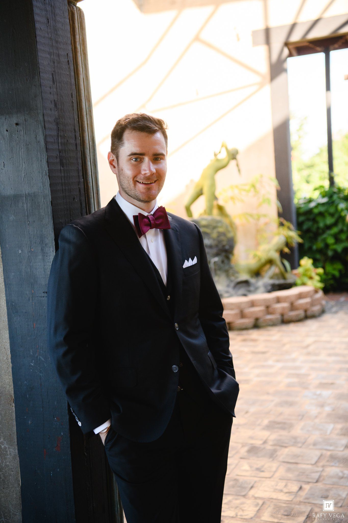 A smiling groom in a black tuxedo with burgundy bow tie and white pocket square poses outdoors on a brick patio.